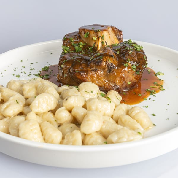 Osso buco with rich brown sauce and fresh parsley, served alongside gnocchi on a white plate.