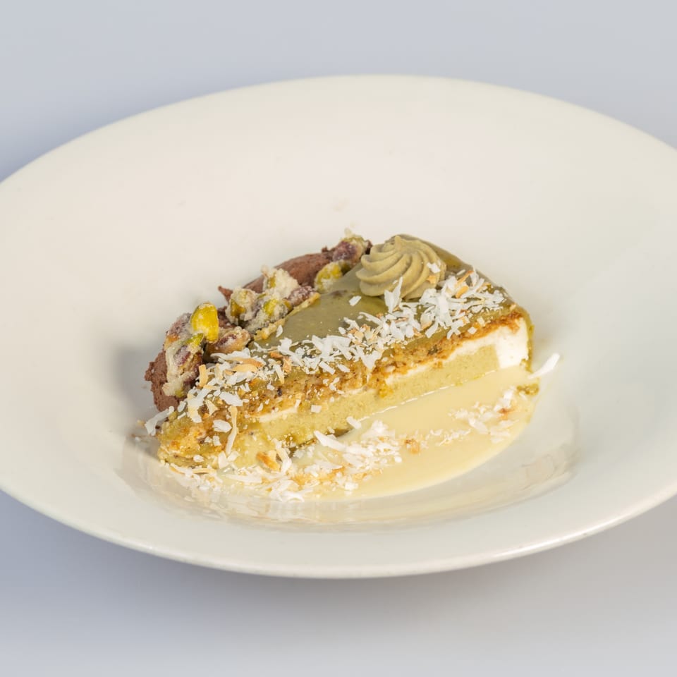 Slice of layered cake with cream, nuts, and shredded coconut on a white plate.