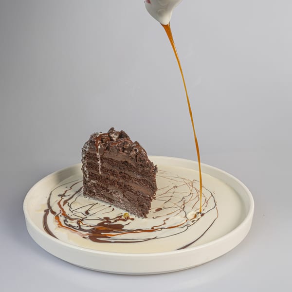 A slice of chocolate cake with caramel sauce being poured over it on a white plate.